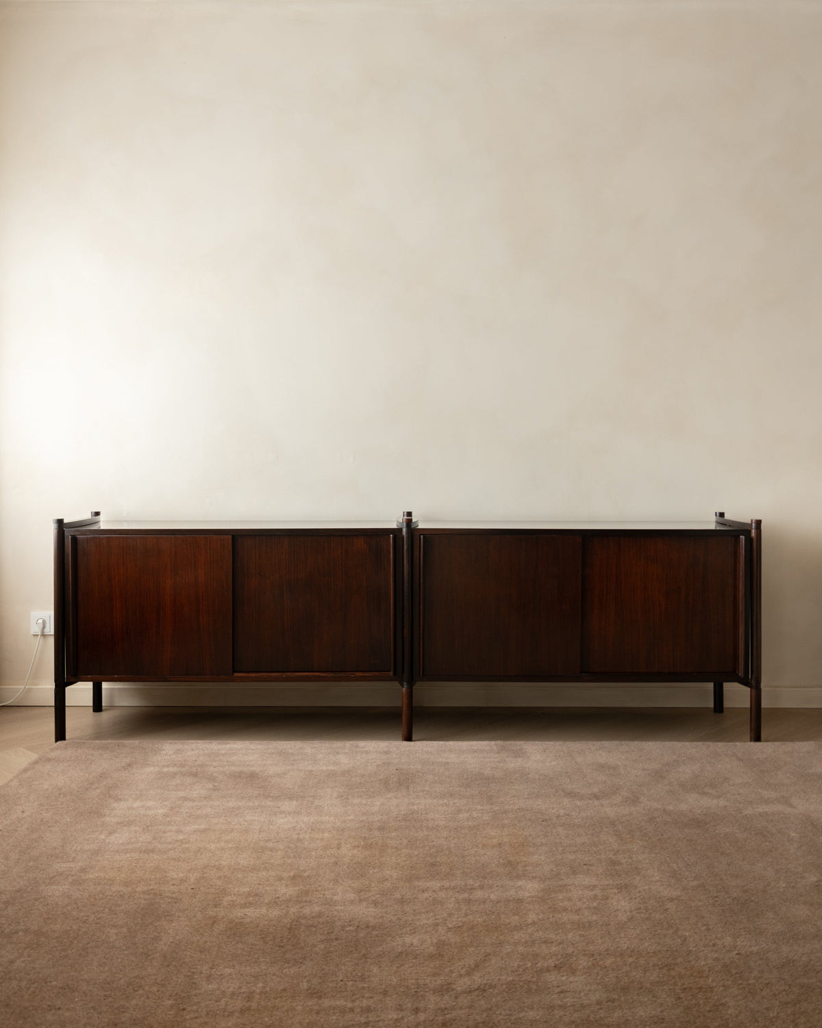A mid-century modern dark wood sideboard cabinet with a warm brown finish, set against a minimalist beige wall and textured brown carpet.