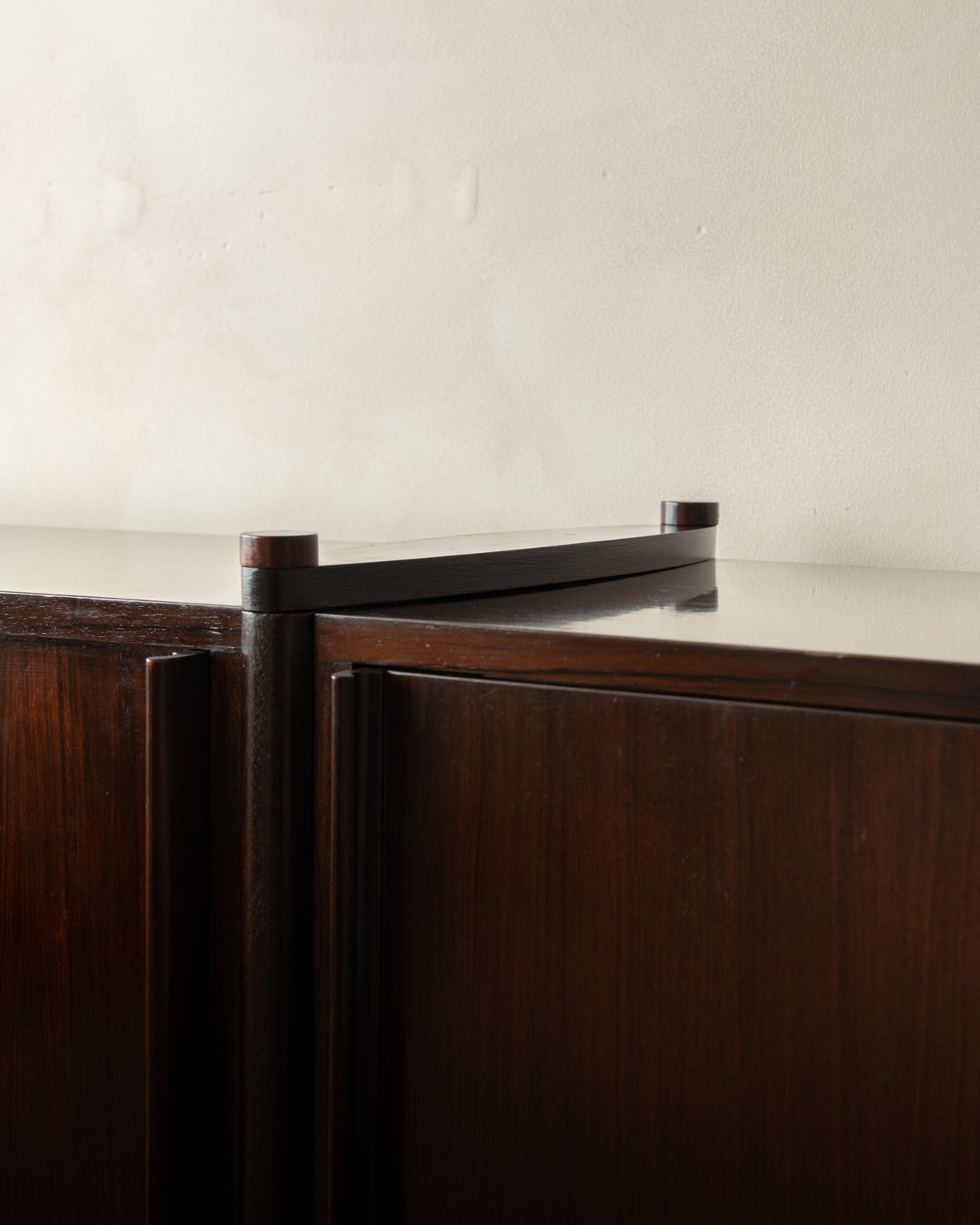 Close-up of a dark wood cabinet with a glossy black metal top rail and cylindrical end caps against a plain off-white wall.