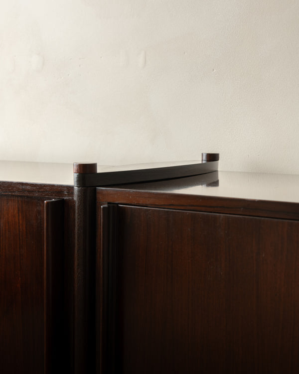 Close-up of a dark wood cabinet with a glossy black metal top rail and cylindrical end caps against a plain off-white wall.