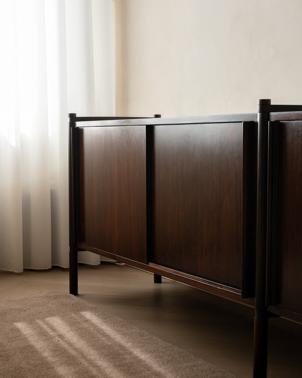 A mid-century modern wooden sideboard with dark brown wood grain doors and a black metal frame, positioned in a room with sheer white curtains and warm natural light.