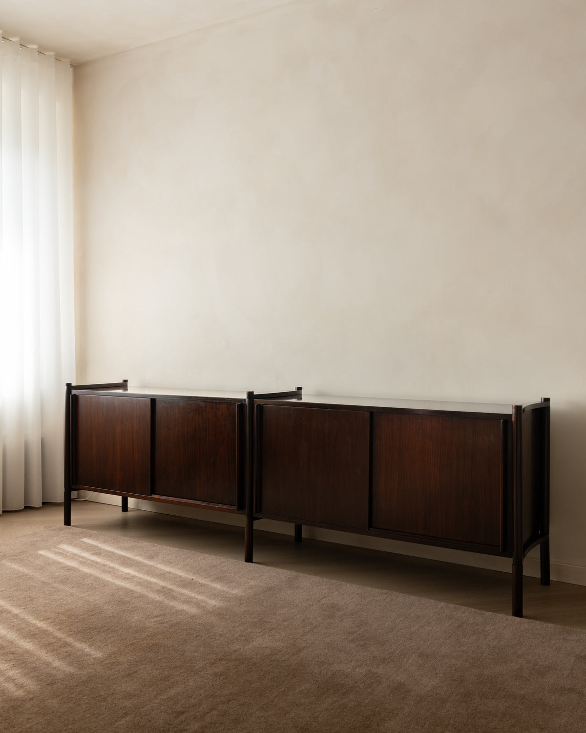 A mid-century modern dark wood credenza with a white marble top, positioned against a textured beige wall in a minimalist room with a beige carpet and sheer white curtains.