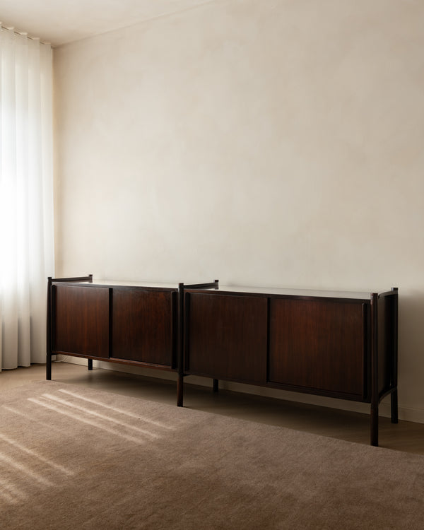 A mid-century modern dark wood credenza with a white marble top, positioned against a textured beige wall in a minimalist room with a beige carpet and sheer white curtains.