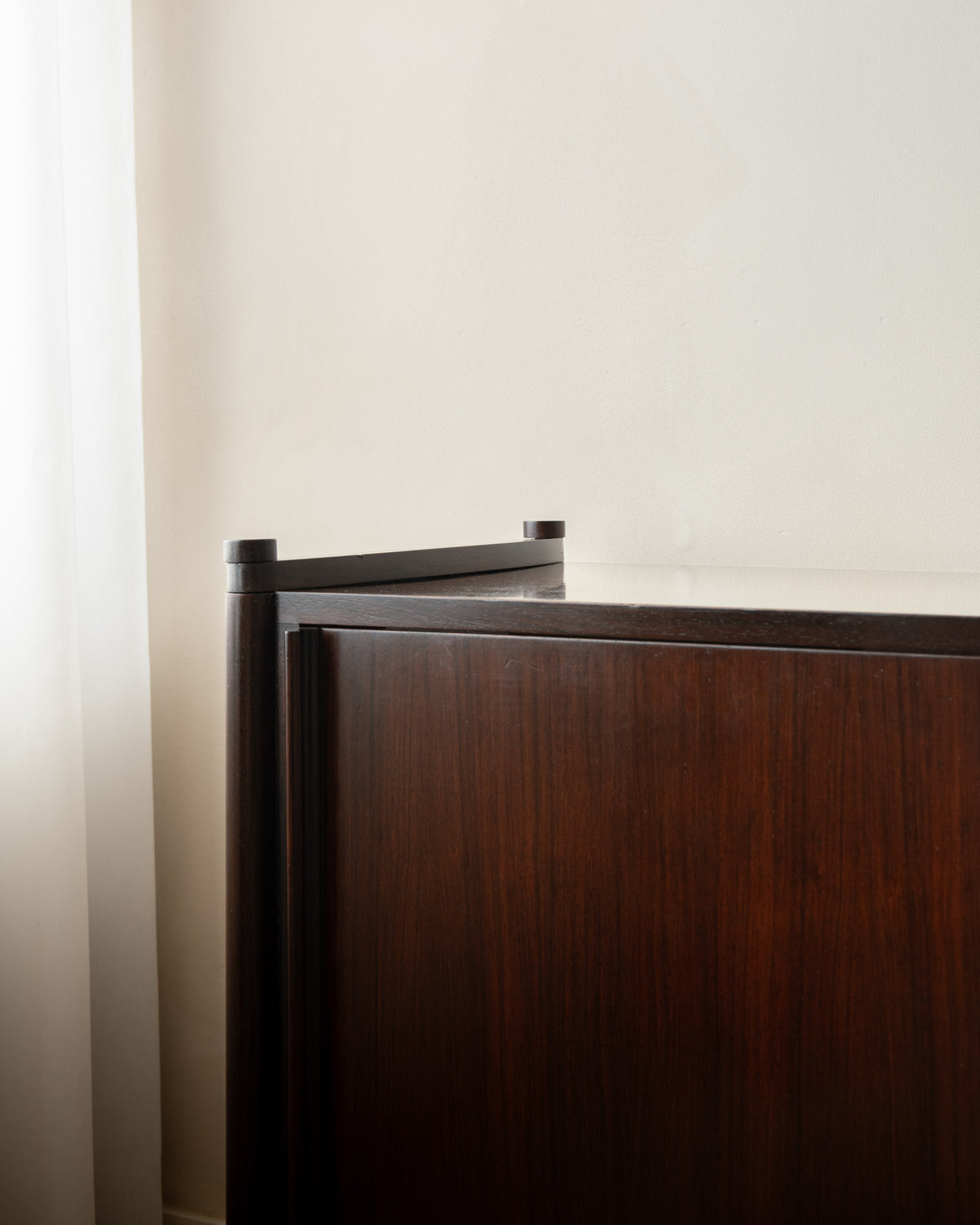 Close-up of a dark brown wooden cabinet or dresser corner with a light-colored wall and a sheer curtain on the left.