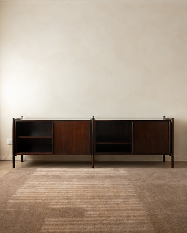 A mid-century modern wooden sideboard cabinet with dark brown wood grain, open shelving, and sliding doors, positioned against a textured beige wall on a striped brown rug.