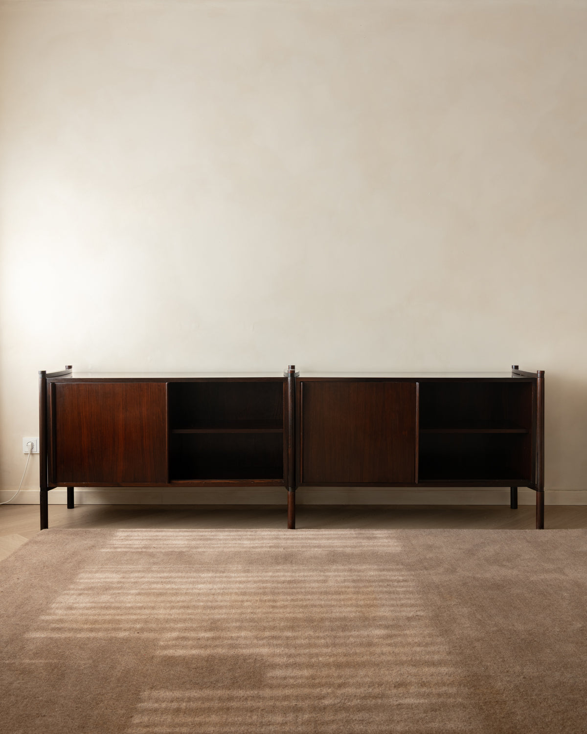 A mid-century modern dark wood sideboard cabinet with open shelving, positioned against a textured beige wall on a neutral brown rug.
