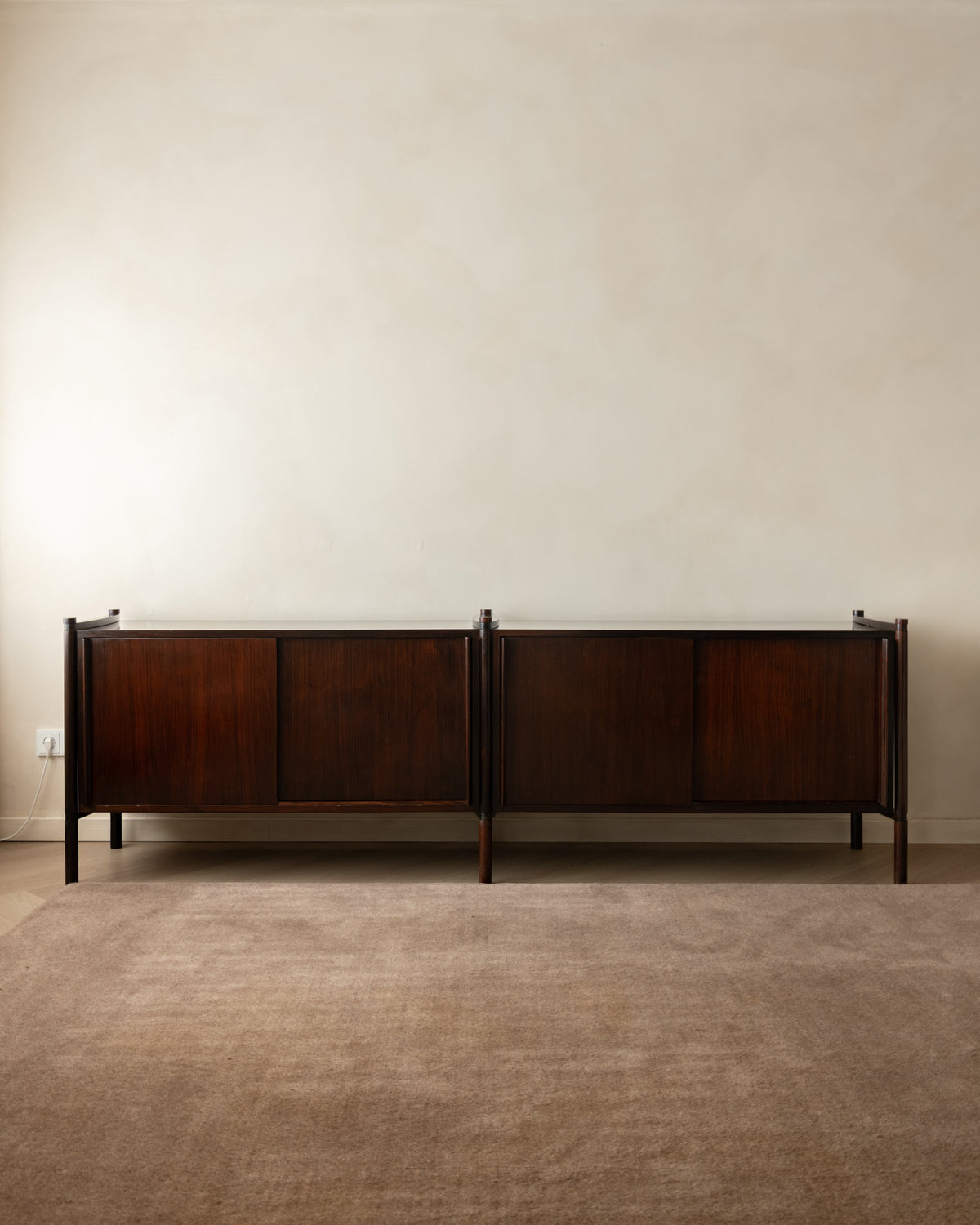 A mid-century modern dark wood sideboard cabinet with a warm brown finish, set against a minimalist beige wall and textured brown carpet.