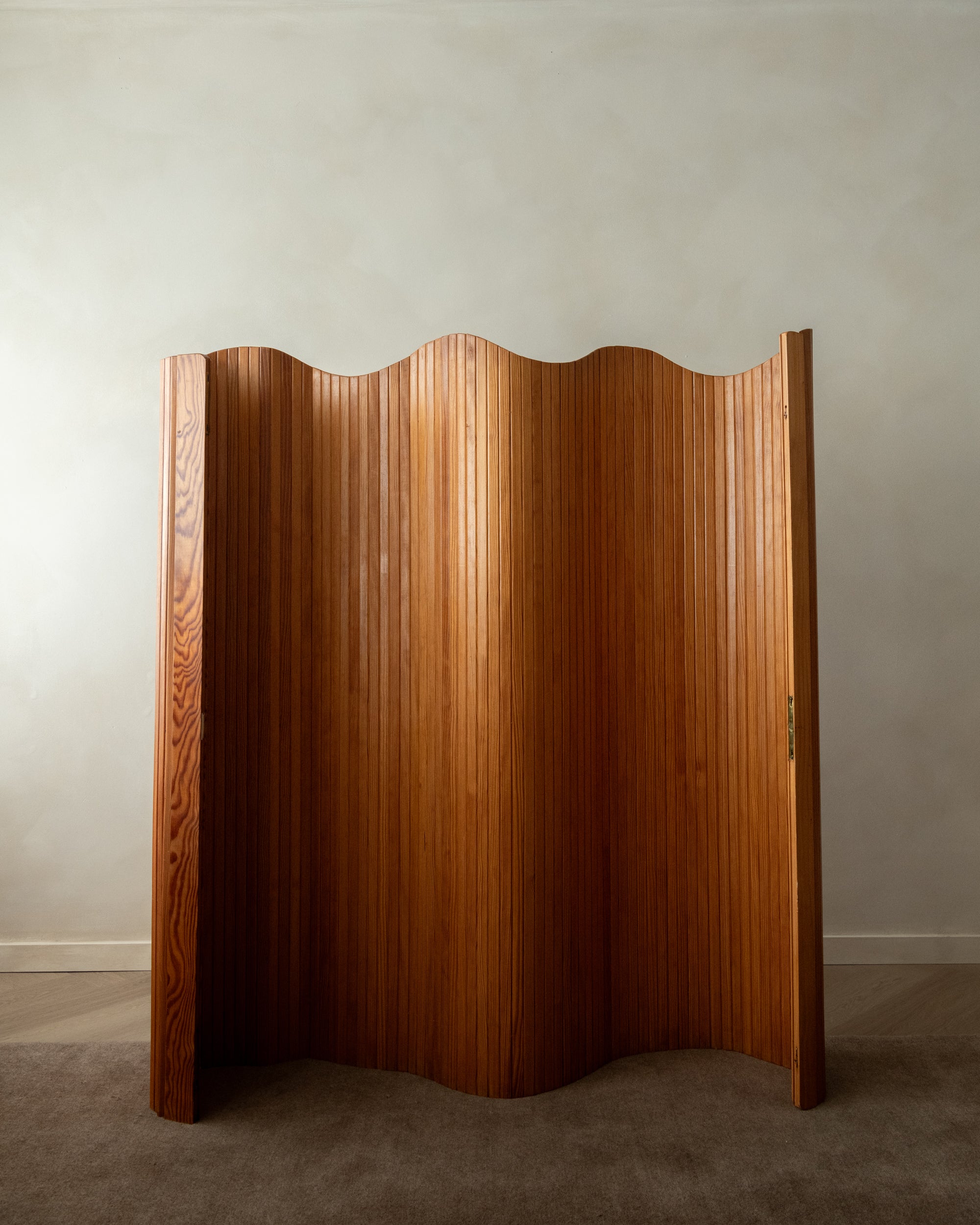 A mid-century modern wooden folding screen with a wavy, pleated design in a warm medium brown finish, standing on a carpeted floor against a neutral gray wall.