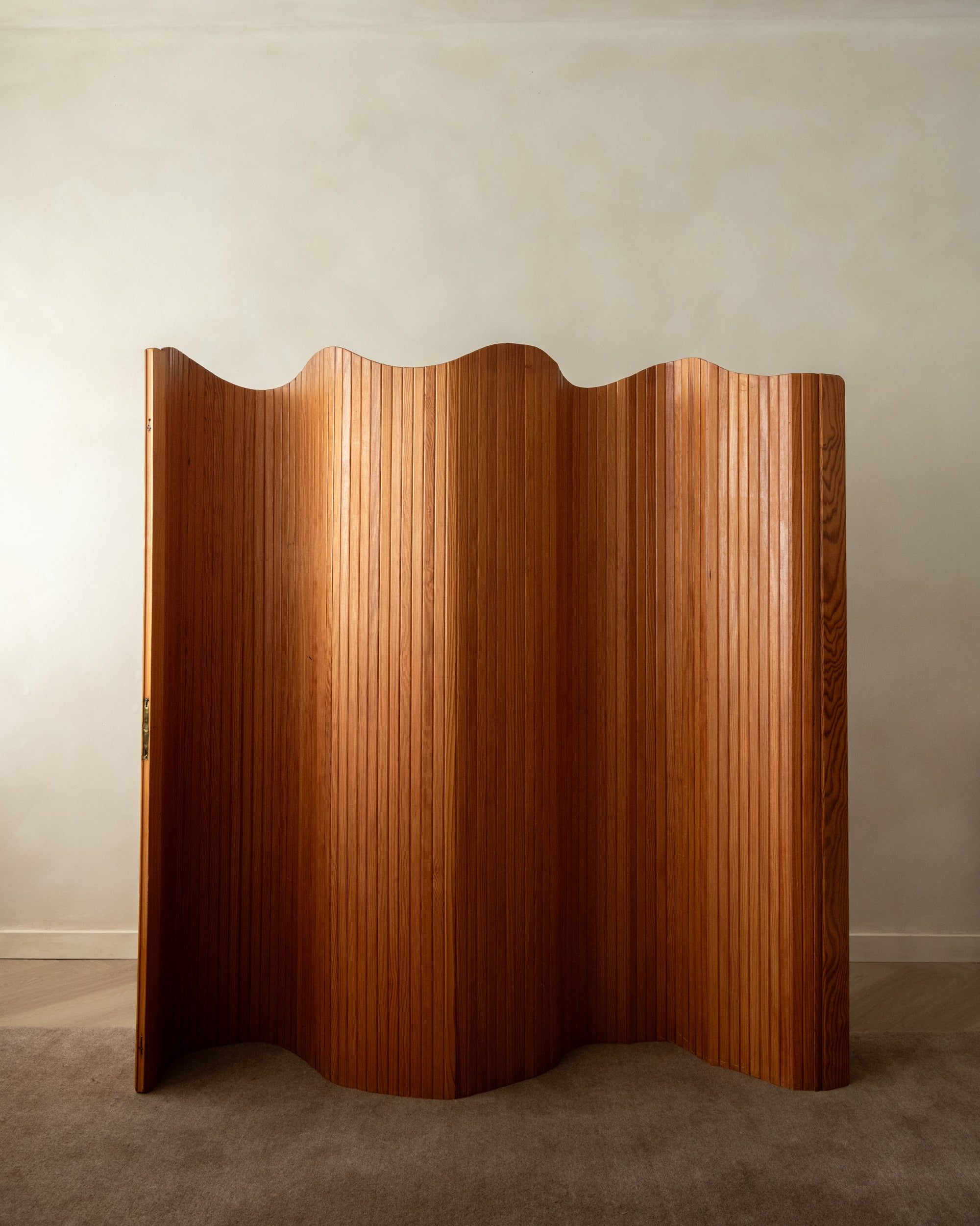 A mid-century modern folding screen made of warm brown wood with a wavy, undulating design, standing on a gray carpet against a neutral beige wall.