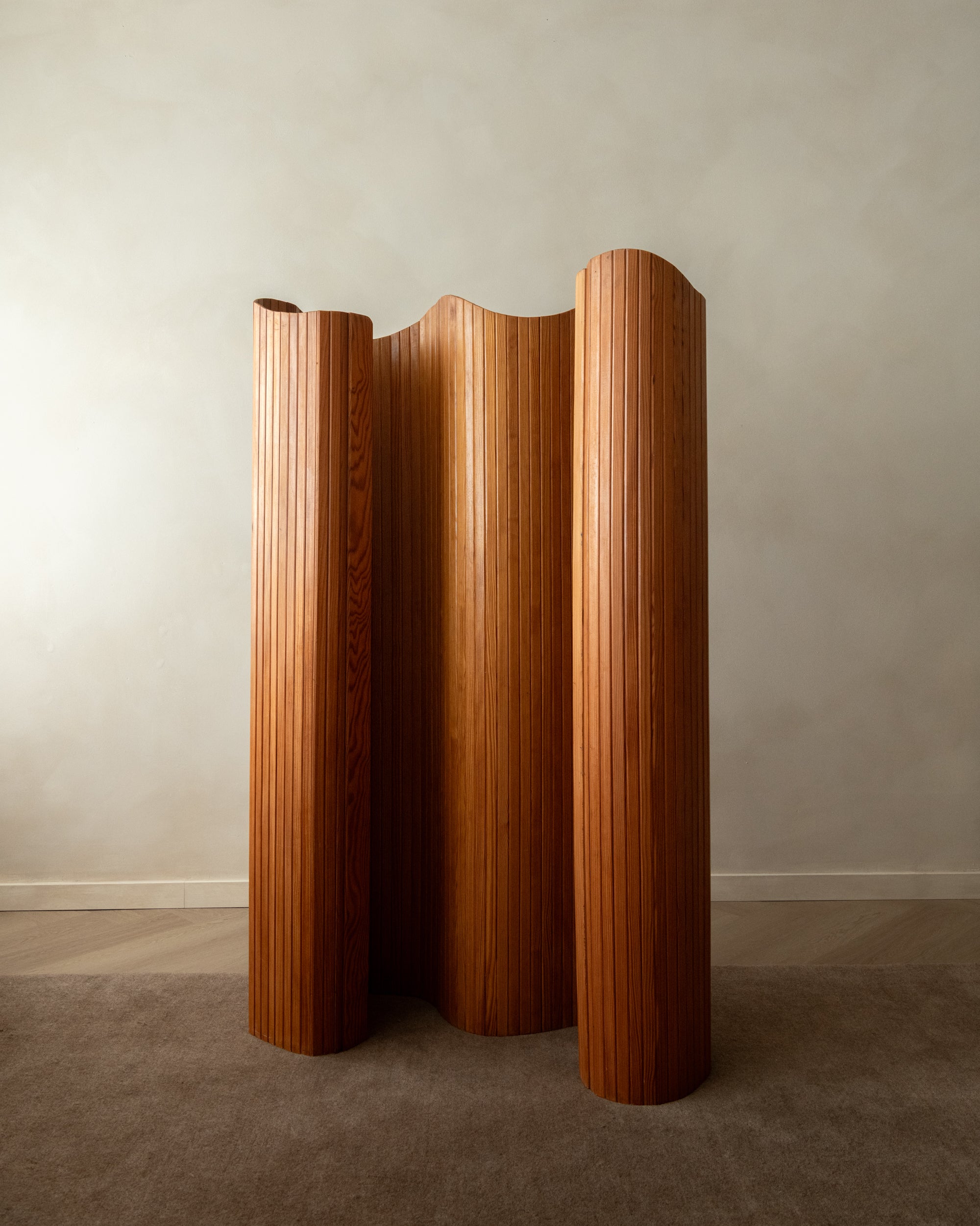 A mid-century modern wooden folding screen with a wavy, pleated design in a warm medium brown finish, standing on a carpeted floor against a neutral gray wall.