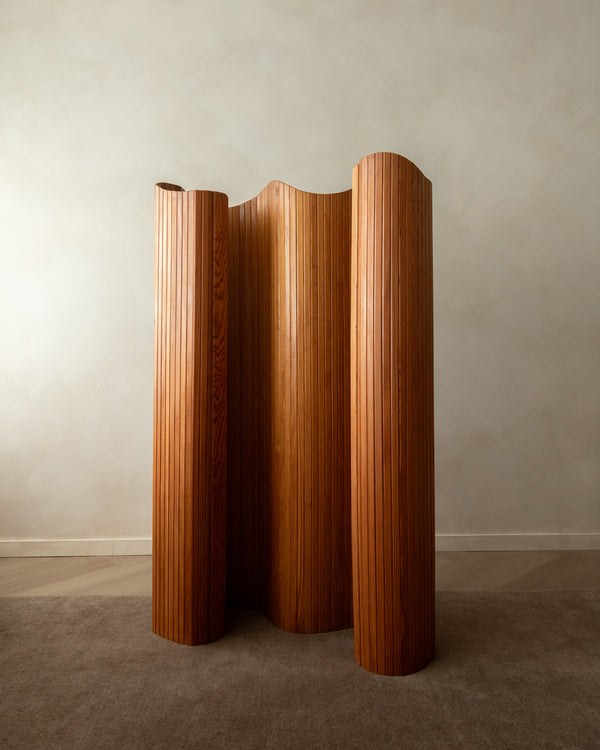 A mid-century modern wooden folding screen with a wavy, pleated design in a warm medium brown finish, standing on a carpeted floor against a neutral gray wall.