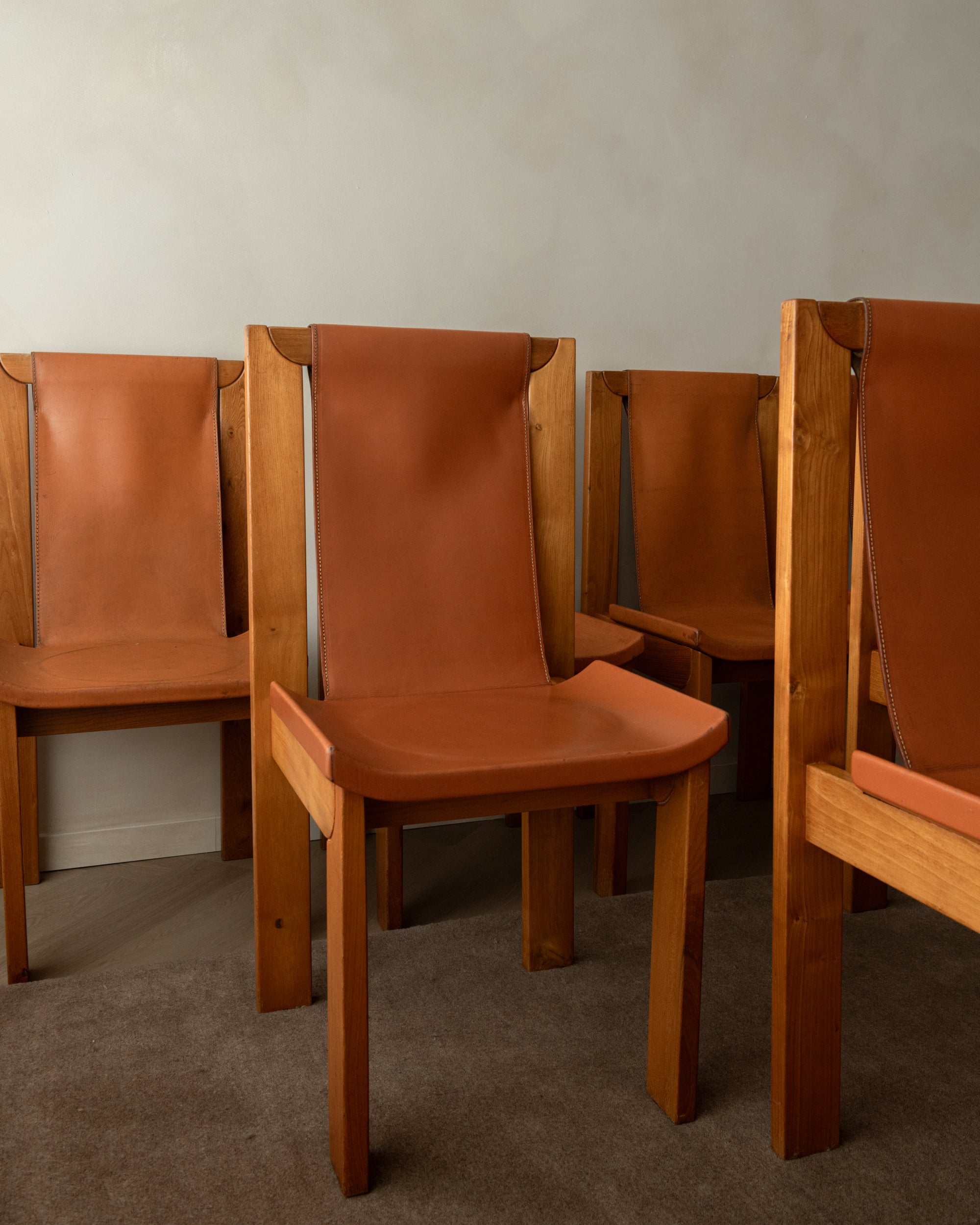 A set of mid-century modern wooden dining chairs with warm brown leather seats and backs, arranged against a plain light gray wall and gray carpet.