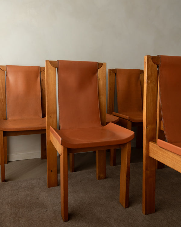A set of mid-century modern wooden dining chairs with warm brown leather seats and backs, arranged against a plain light gray wall and gray carpet.