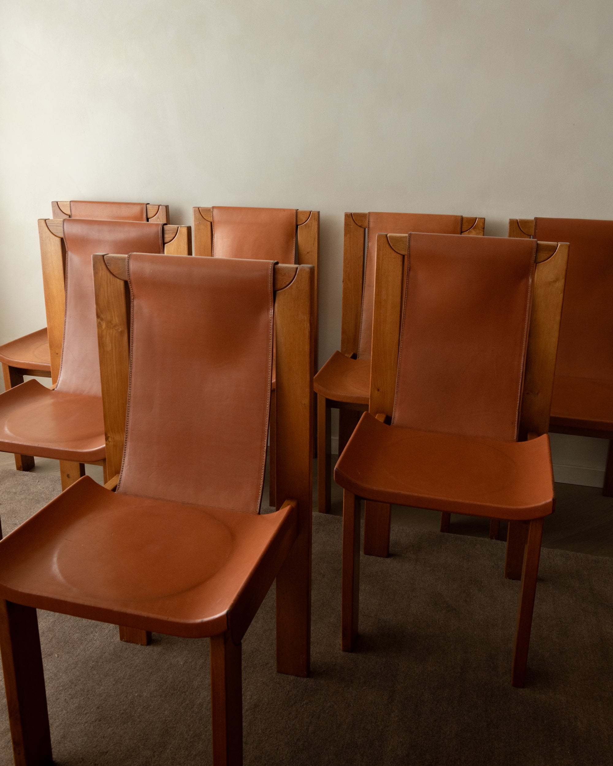 A group of mid-century modern wooden dining chairs with curved brown leather seats and tall wooden backs, arranged against a plain off-white wall.