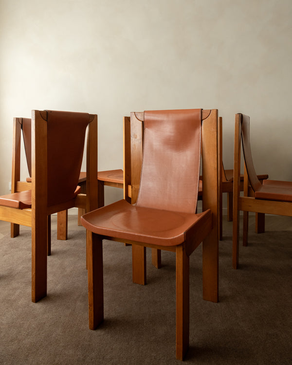 A mid-century modern wooden dining chair with a curved brown leather seat and backrest, shown in a room setting with other matching chairs on a carpeted floor against a textured beige wall.