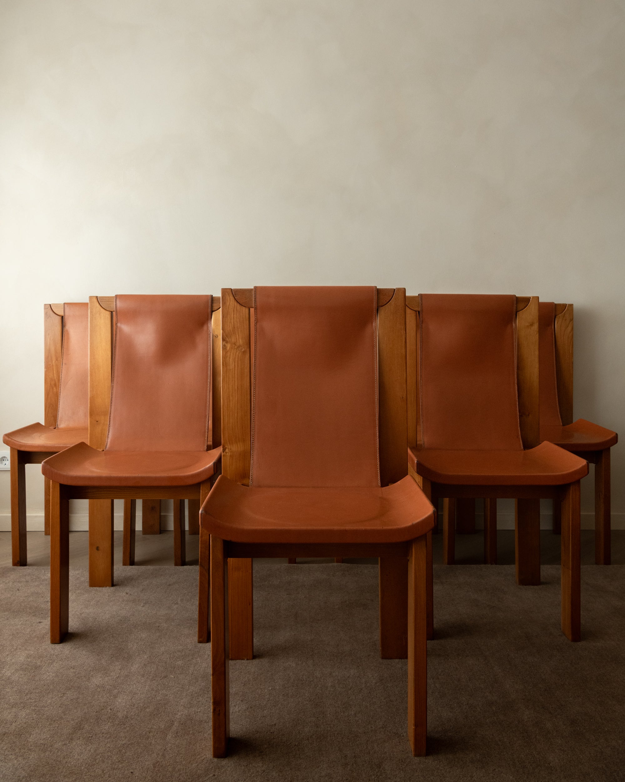 A set of mid-century modern wooden dining chairs with curved tan leather seats and tall, straight backs, arranged against a neutral beige wall and textured gray carpet.
