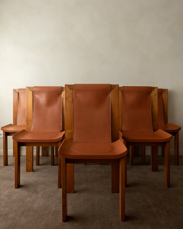 A set of mid-century modern wooden dining chairs with curved tan leather seats and tall, straight backs, arranged against a neutral beige wall and textured gray carpet.