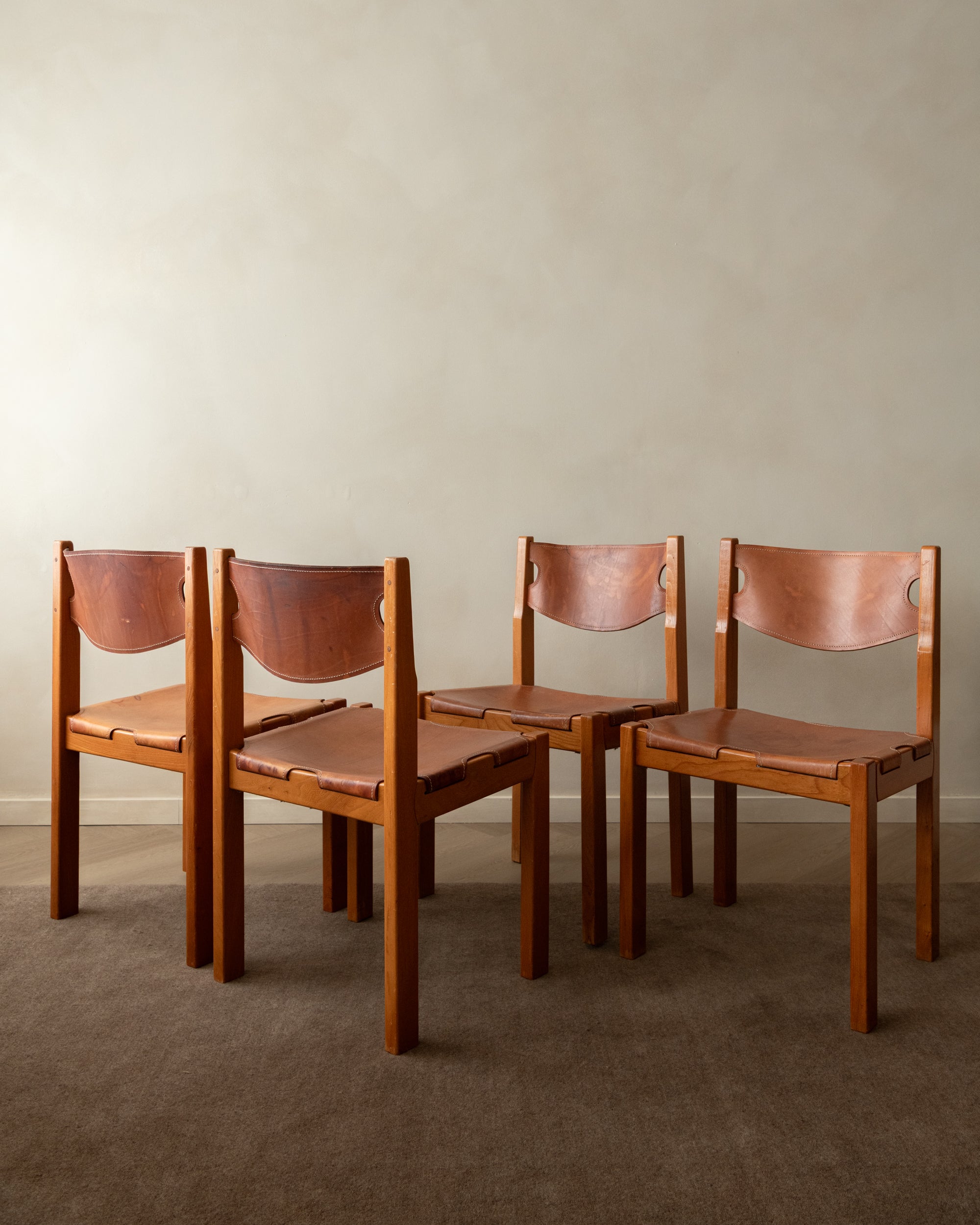 Four mid-century modern wooden dining chairs with curved copper-brown leather seats and backs, arranged on a brown carpet against a neutral beige wall.