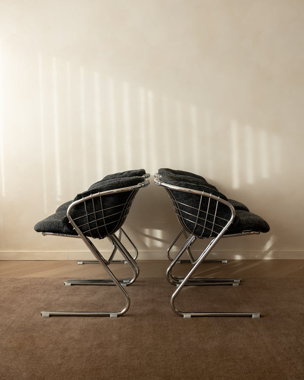 Two mid-century modern wire chairs with dark gray cushions and chrome tubular steel frames, positioned side-by-side on a brown carpeted floor against a beige wall with soft window shadow patterns.