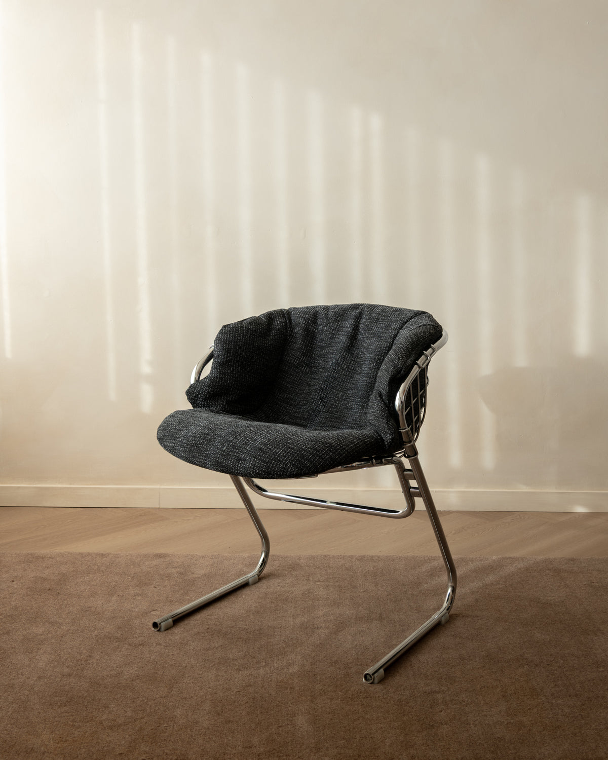A modern accent chair with a dark gray textured fabric seat and curved chrome metal legs, positioned on a brown carpet against a beige wall with soft vertical shadows.