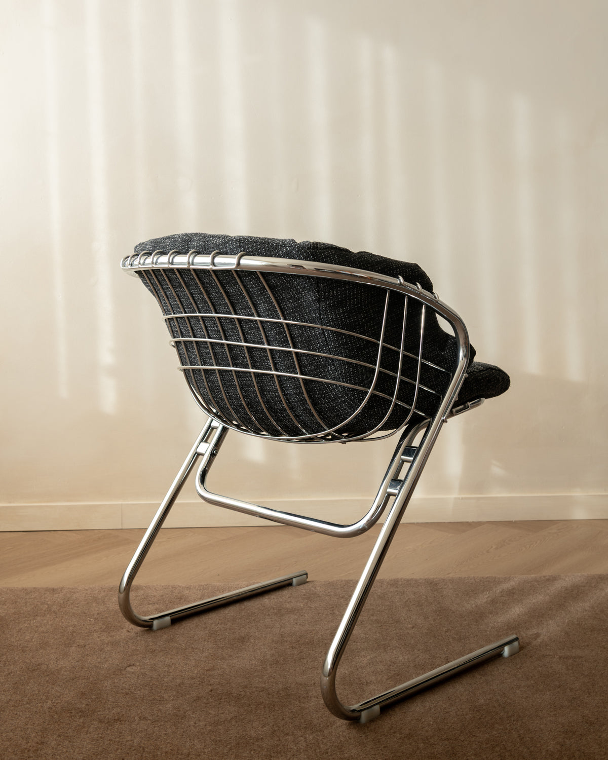 A mid-century modern wire chair with a black woven seat and backrest, supported by a chrome cantilever frame, set on a brown carpet against a beige wall with soft window shadow patterns.