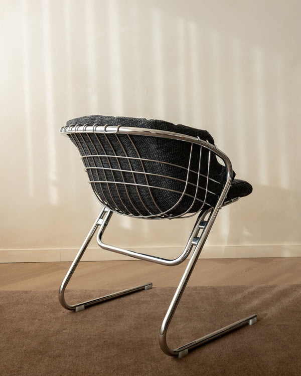 A mid-century modern wire chair with a black woven seat and backrest, supported by a chrome cantilever frame, set on a brown carpet against a beige wall with soft window shadow patterns.