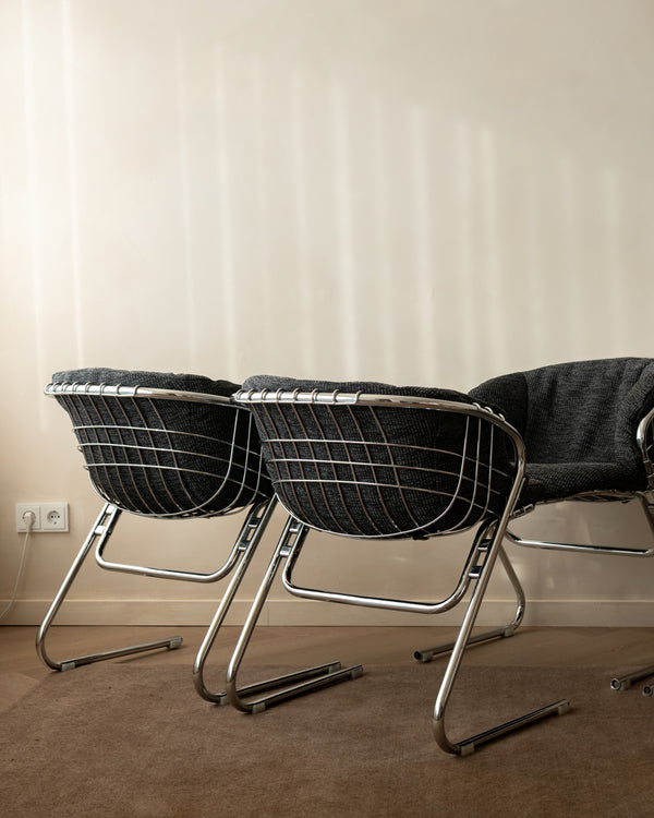 A pair of modern wire-frame dining chairs with dark gray textured cushions and chrome cantilever legs, positioned against a warm beige wall with soft window light shadows.