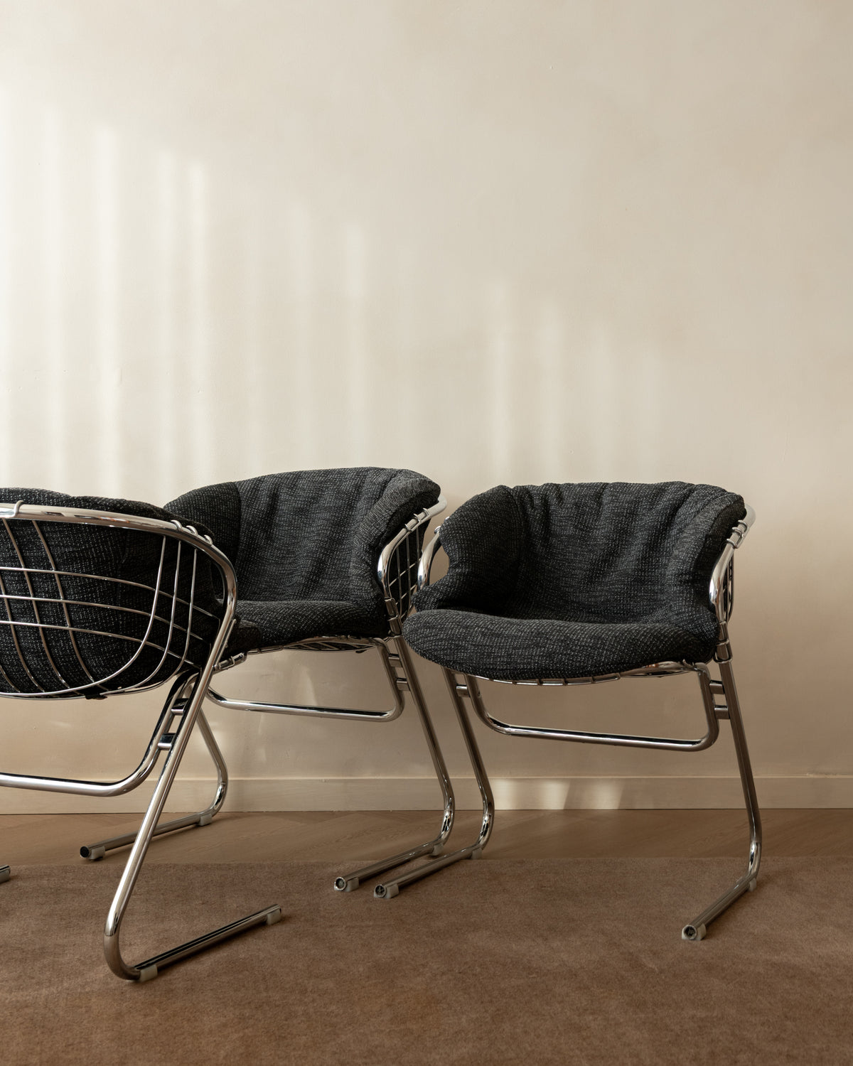 Two modern accent chairs with dark gray textured fabric seats and chrome wire and metal frame legs, set against a warm beige wall and brown carpet.