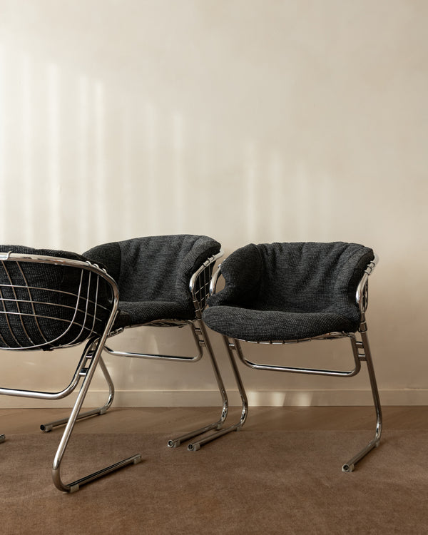 Two modern accent chairs with dark gray textured fabric seats and chrome wire and metal frame legs, set against a warm beige wall and brown carpet.