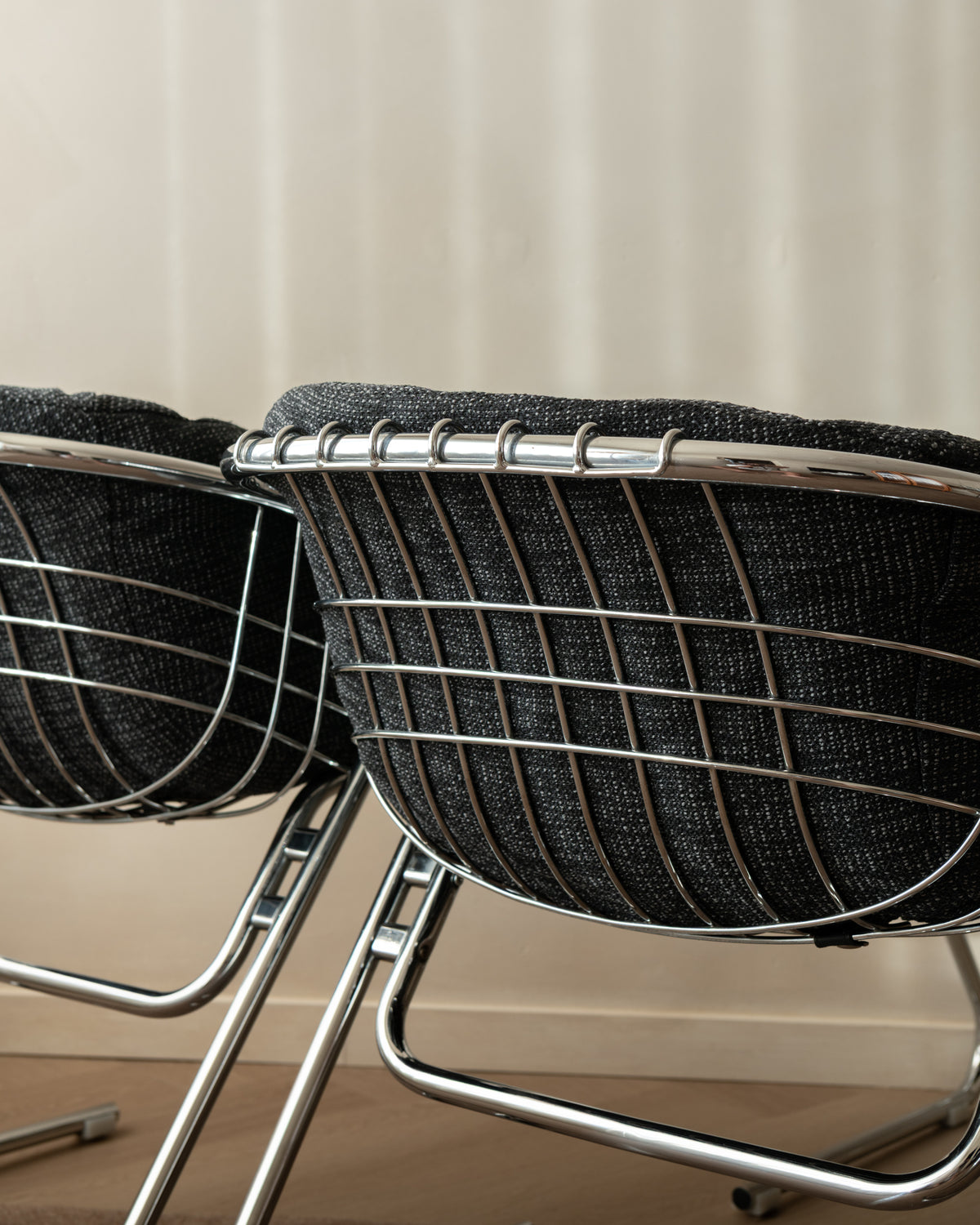 A close-up of a modern wire chair with a dark charcoal woven seat cushion and polished chrome metal frame, set against a neutral beige background.