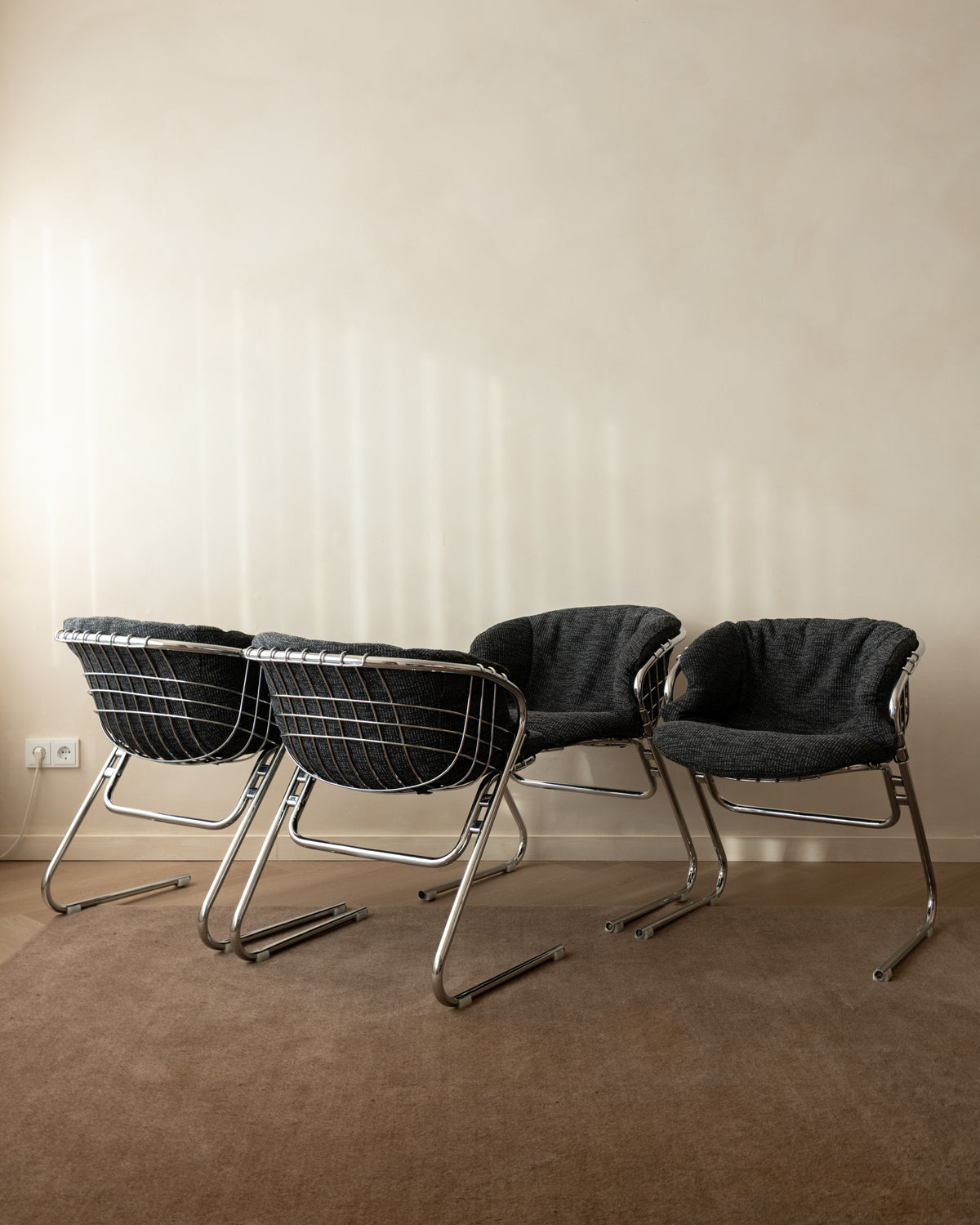 Three modern lounge chairs with dark gray woven seats and chrome tubular metal frames, arranged on a brown carpet against a beige wall.