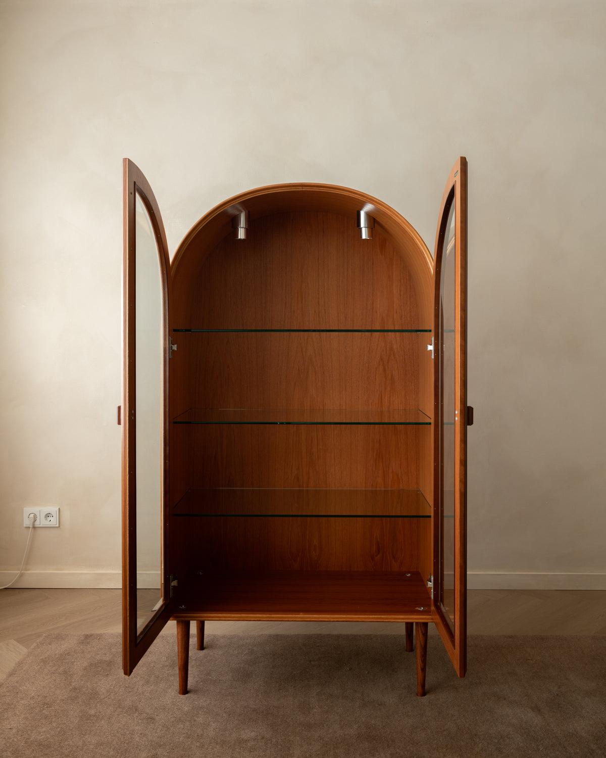 A mid-century modern wooden display cabinet with an arched top, glass doors, and three glass shelves, shown open against a neutral beige wall and carpeted floor.