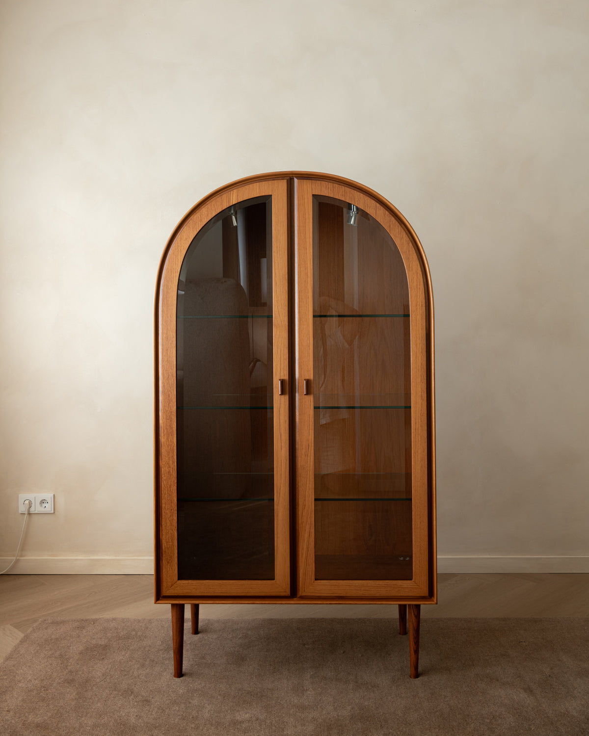 Mid-century modern wooden display cabinet with arched double glass doors, glass shelves, and tapered legs, set against a neutral beige wall and carpet.