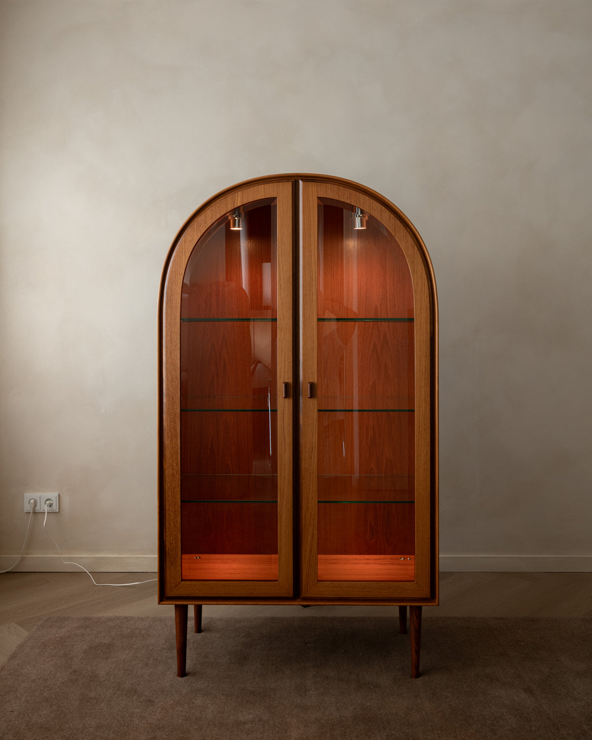 An arched wooden display cabinet with glass doors and interior lighting, set against a plain beige wall and carpeted floor.