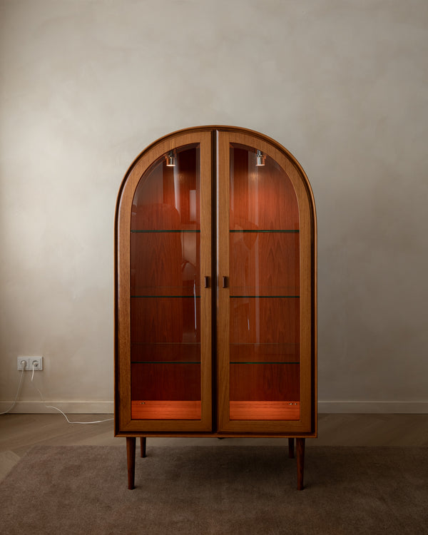 An arched wooden display cabinet with glass doors and interior lighting, set against a plain beige wall and carpeted floor.