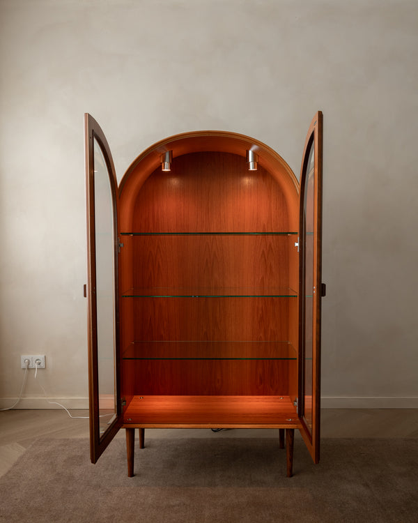 A mid-century modern wooden display cabinet with an arched back panel, glass doors, and four glass shelves, shown open against a neutral beige wall.