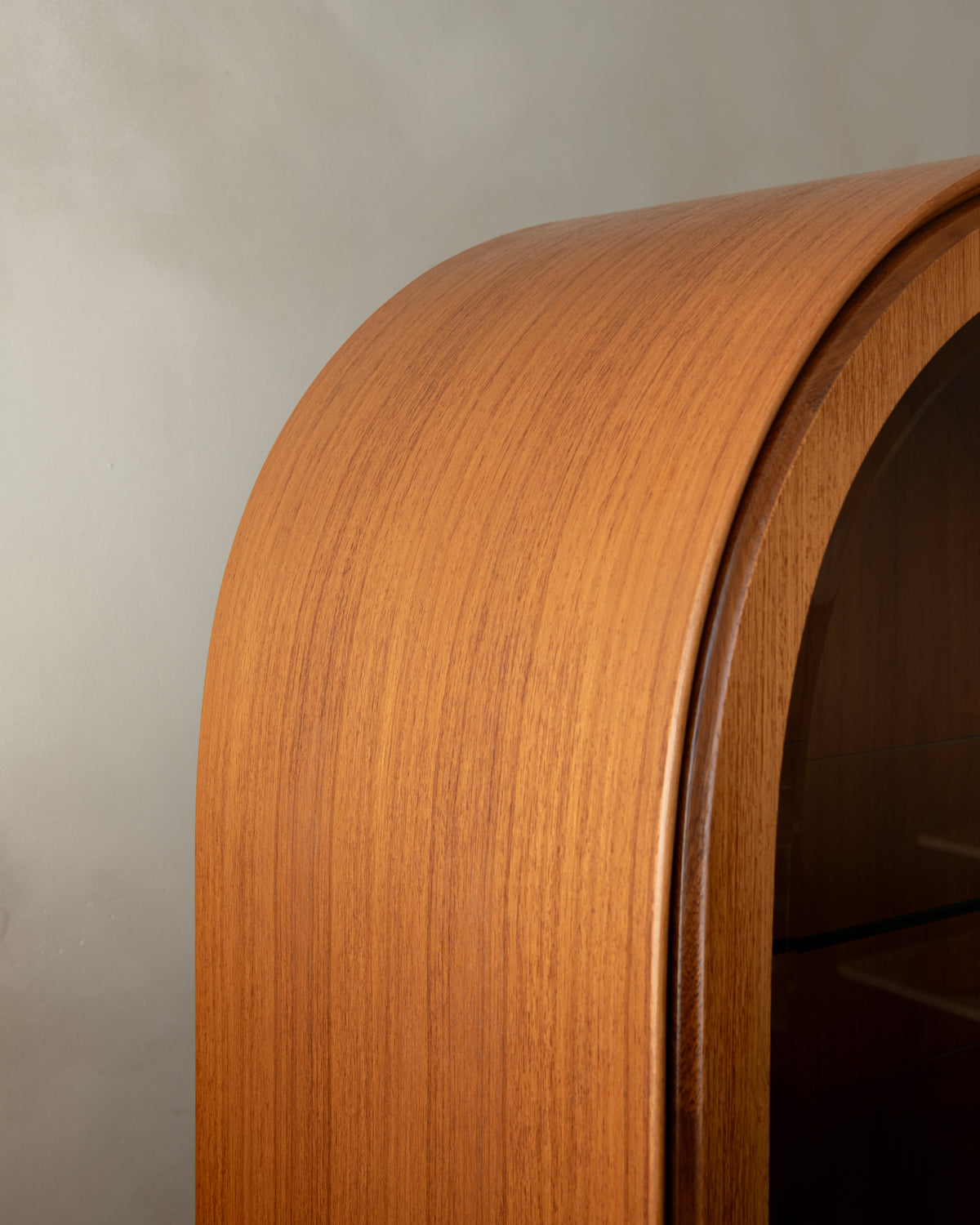 Close-up of a curved wooden cabinet or shelf with a warm honey-toned wood grain finish against a neutral gray wall.