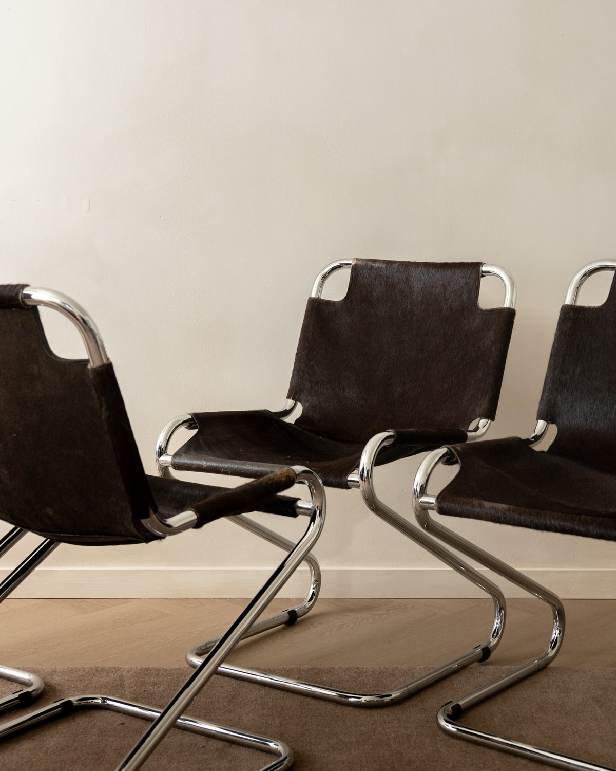 Three modern cantilever chairs with dark brown leather seats and curved chrome metal frames, arranged on a carpeted floor against a plain beige wall.