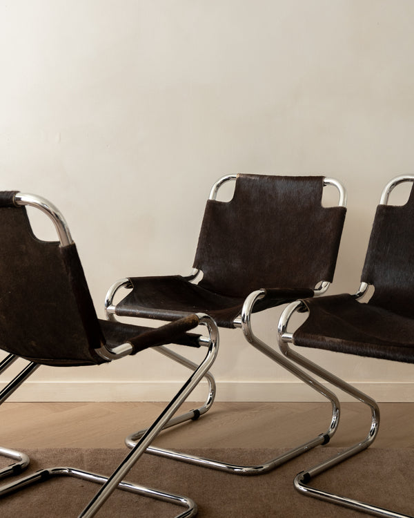 Three modern cantilever chairs with dark brown leather seats and curved chrome metal frames, arranged on a carpeted floor against a plain beige wall.