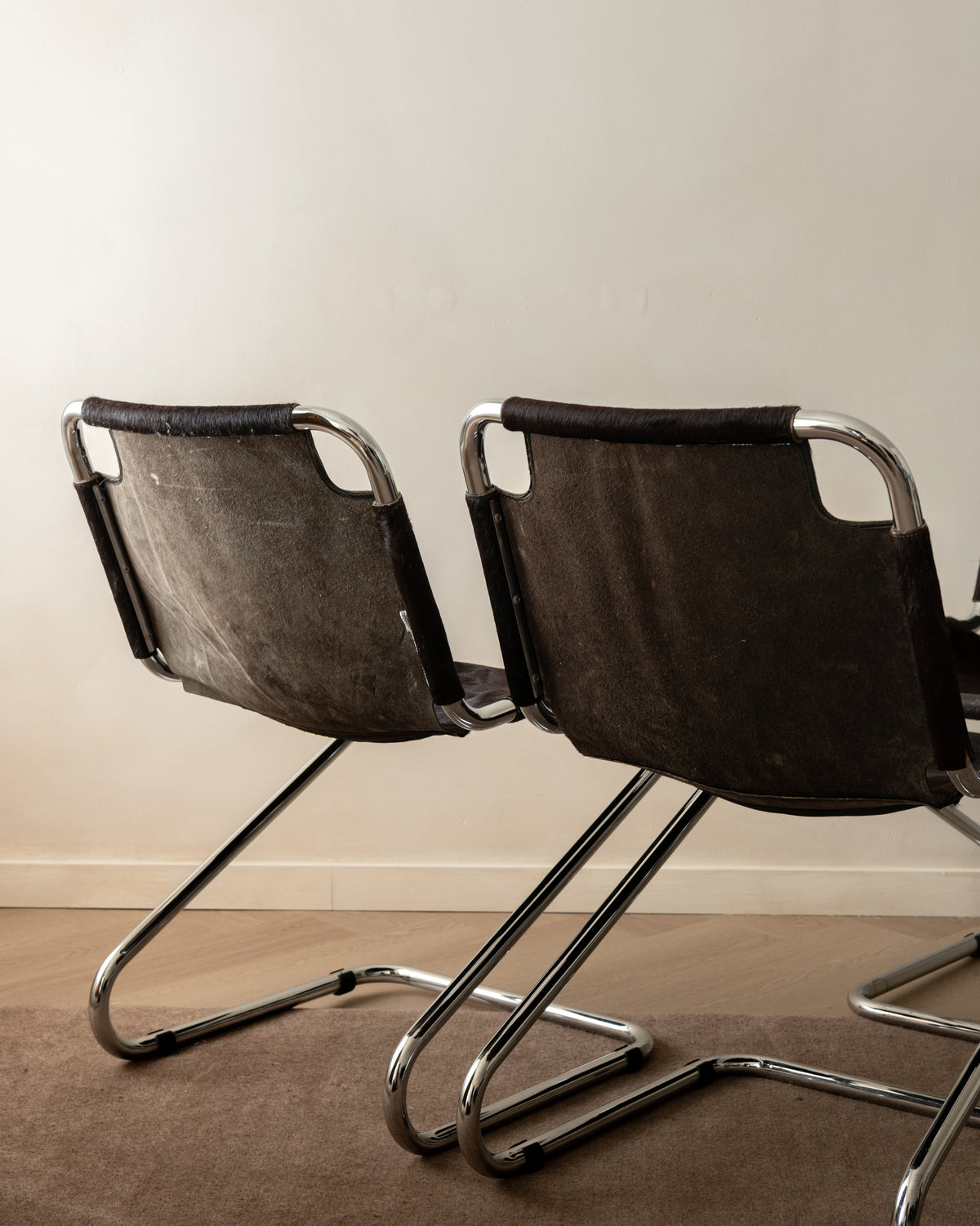 Two modern cantilever chairs with dark brown textured fabric seats and chrome metal frames, positioned side-by-side against a plain beige wall.