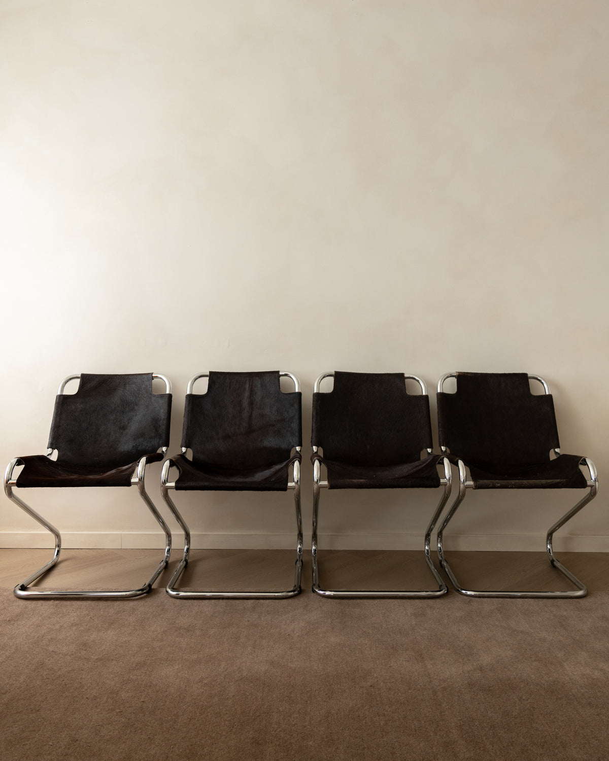 Four black canvas camp chairs with chrome tubular steel frames lined up against a plain beige wall and carpeted floor.