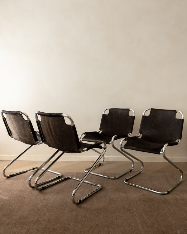 Four modern cantilever chairs with dark brown textured leather seats and backs and chrome tubular steel frames, arranged in a row on a brown carpet against a beige wall.