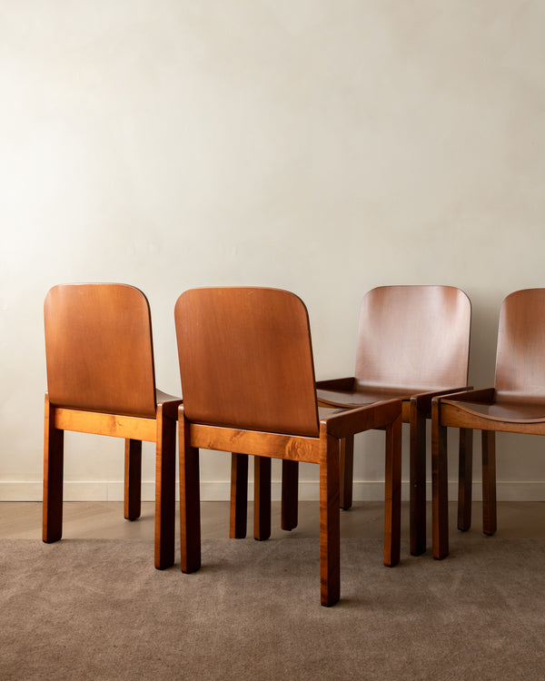 Four mid-century modern wooden dining chairs with curved tan leather seats and backs, arranged against a neutral beige wall and textured brown carpet.
