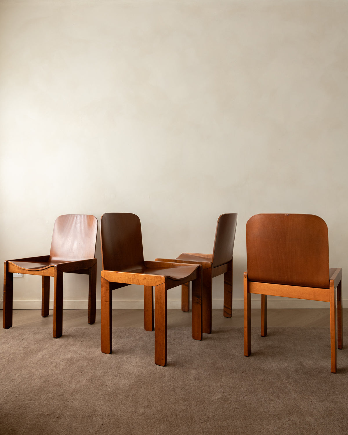 Four mid-century modern wooden chairs in various shades of brown, arranged on a textured gray carpet against a warm beige plaster wall.
