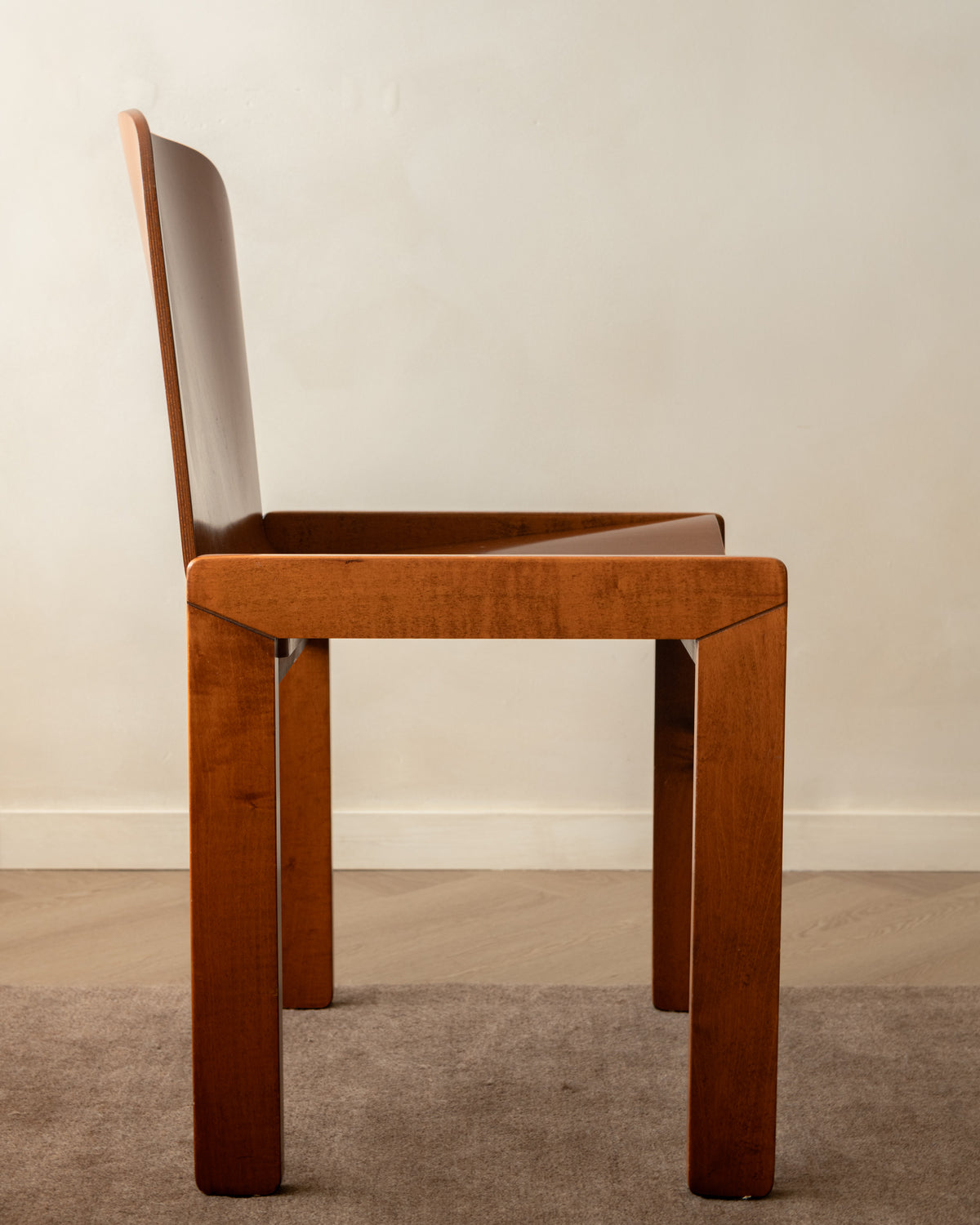 A mid-century modern wooden dining chair with a warm brown finish, shown from a side profile against a neutral beige wall and gray carpet.
