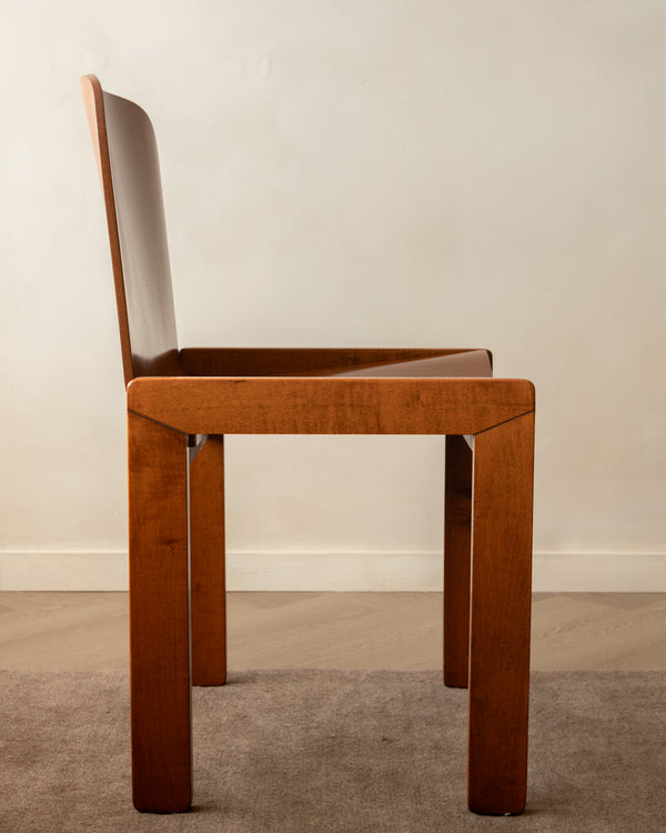 A mid-century modern wooden dining chair with a warm brown finish, shown from a side profile against a neutral beige wall and gray carpet.