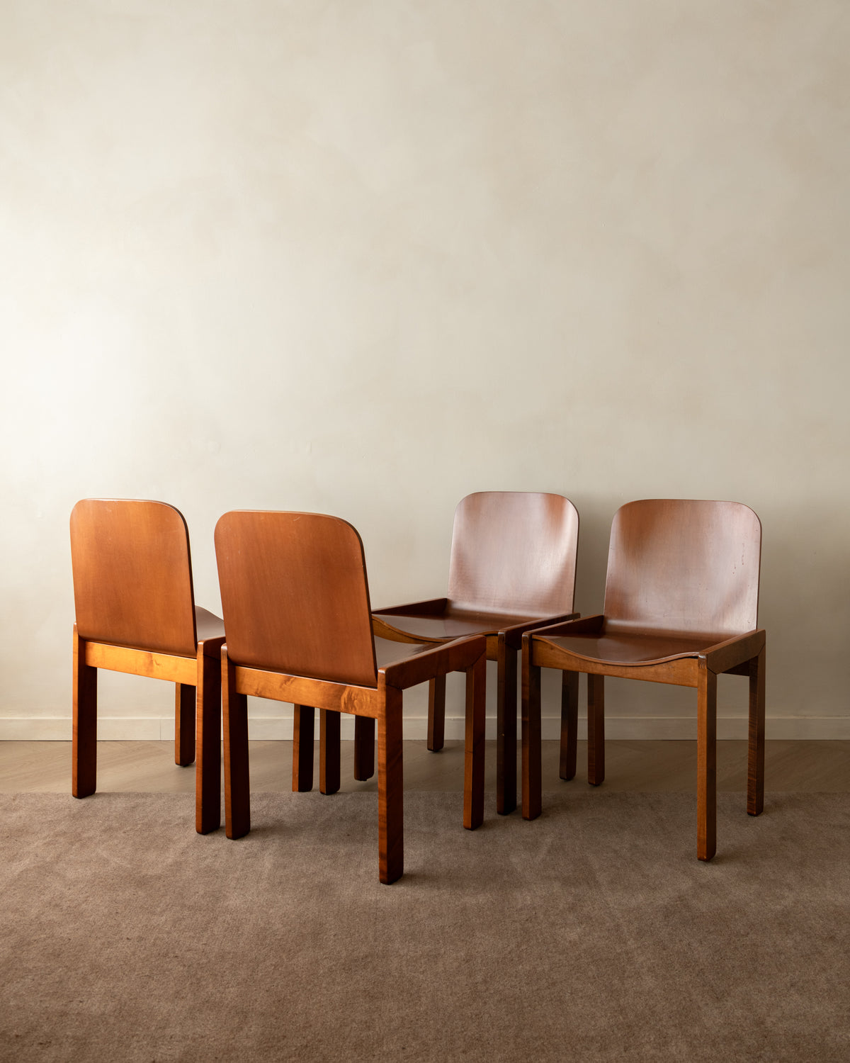 Four mid-century modern dining chairs with warm brown wood frames and smooth tan leather seats, arranged in a semi-circle against a neutral beige wall and carpeted floor.