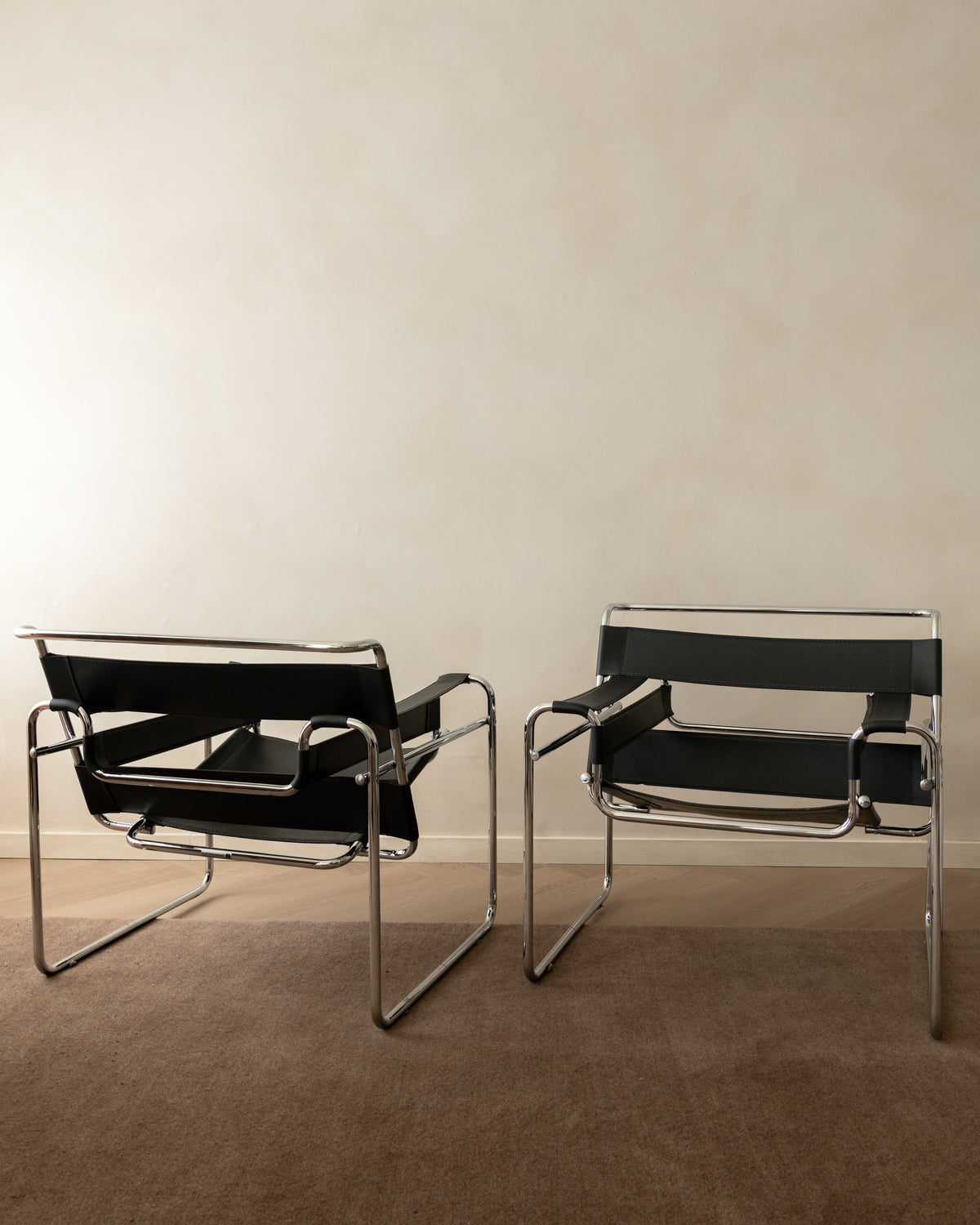 Two classic Wassily armchairs with black leather straps and chrome tubular steel frames, positioned side-by-side on a brown carpeted floor against a neutral beige wall.