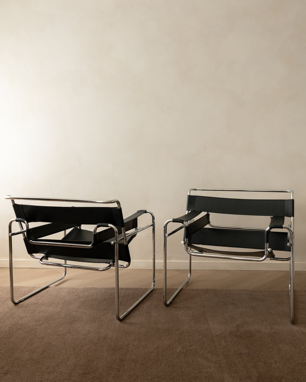 Two classic Wassily armchairs with black leather straps and chrome tubular steel frames, positioned side-by-side on a brown carpeted floor against a neutral beige wall.