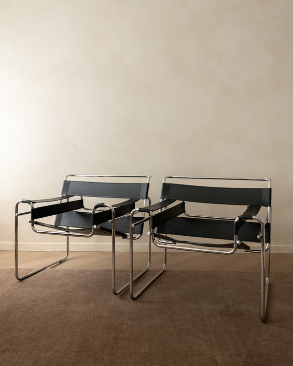Two modern metal-framed chairs with black sling seats and backs, positioned side by side against a plain beige wall and brown carpet.