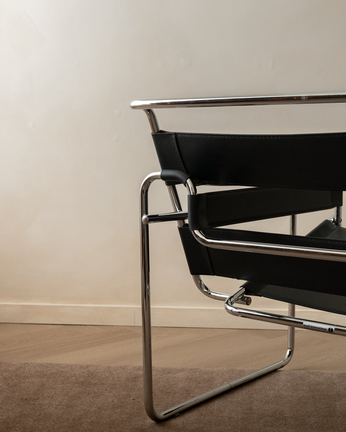 A close-up of a Wassily chair with a black leather sling seat and back, supported by a polished chrome tubular steel frame, set against a neutral beige wall and light wood floor.